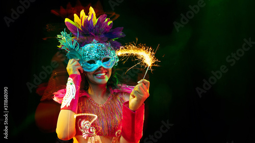 Beautiful young woman in carnival mask, stylish masquerade costume with feathers and sparklers inviting. Flyer for ad on black background. Christmas, New Year, celebration. Festive time, dance, party.