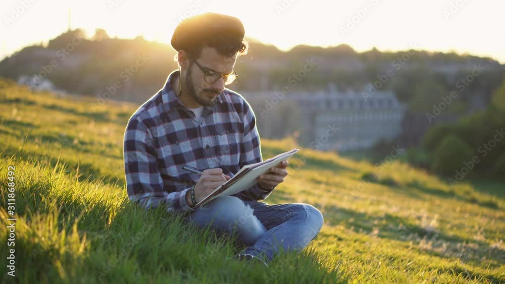 Handsome Young Artist Sketching and Drawing Landscape While Sitting in Grass During Colorful Sunset Handsome Young Artist Sketching and Drawing Landscape While Sitting in Grass During Colorful Sunset