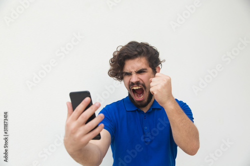 Angry furious guy with smartphone reading message. Handsome bearded young man in blue casual t-shirt posing isolated over white background. Bad news concept