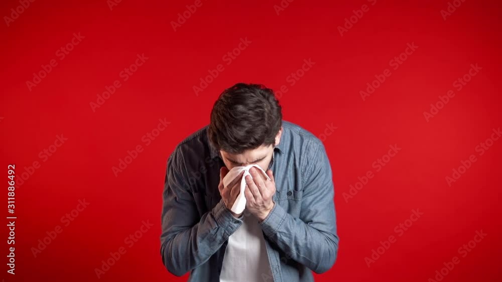 Young man sneezes into tissue. Isolated guy on red studio background is sick, has a cold or allergic reaction. Health, medicine, illness, treatment concept. Young man sneezes into tissue. Isolated guy on red studio background is sick, has a cold or allergic reaction. Health, medicine, illness, treatment concept.