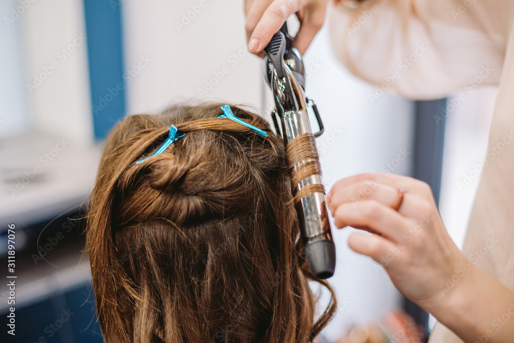 Fototapeta premium beauty, hairstyle concept, happy young woman and hairdresser with hair iron making hairdo at hair salon. Woman Having Hair Stylized Hairdresser. Smoothes Hair Curling. Stylist Using Tool for Modeling
