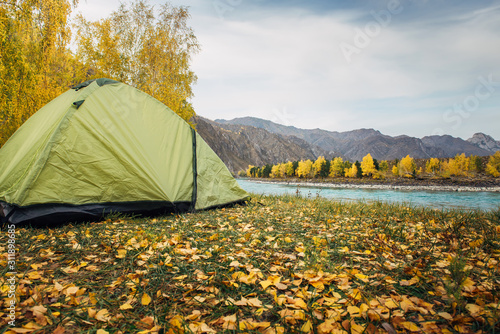 Tourist tent on the grass in a picturesque secluded place on the river bank among rocky mountains on warm autumn day  close-up. Equipment for hiking.