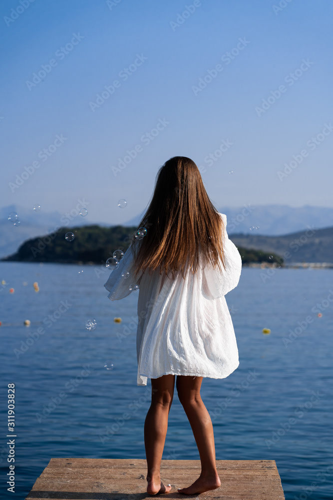 Young beautiful woman in white dress and sunglasses blowing soap bubbles on pier with seaview background. The concept of joy, ease and freedom during the vacation. The girl is enjoying the rest. Young beautiful woman in white dress and sunglasses blowing soap bubbles on pier with seaview background. The concept of joy, ease and freedom during the vacation. The girl is enjoying the rest.