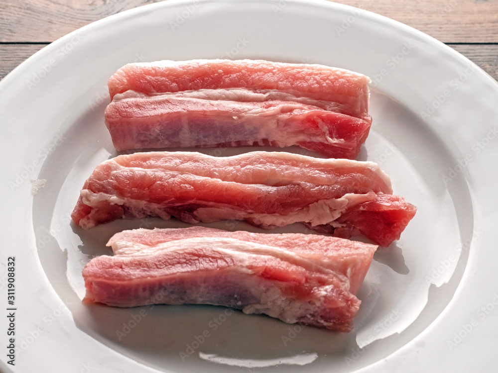 Uncooked raw pork belly slices on a white plate and wooden table. Uncooked raw pork belly slices on a white plate and wooden table.