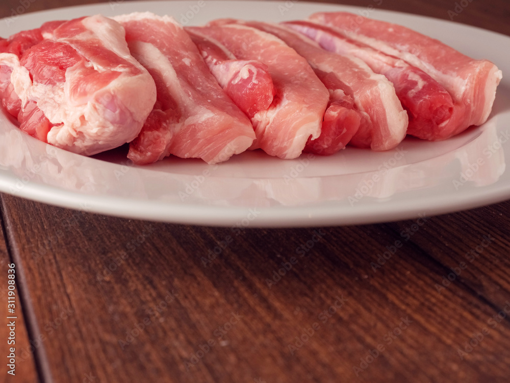 Fresh uncooked pork belly slices on a white plate. Wooden table surface. Fresh uncooked pork belly slices on a white plate. Wooden table surface.