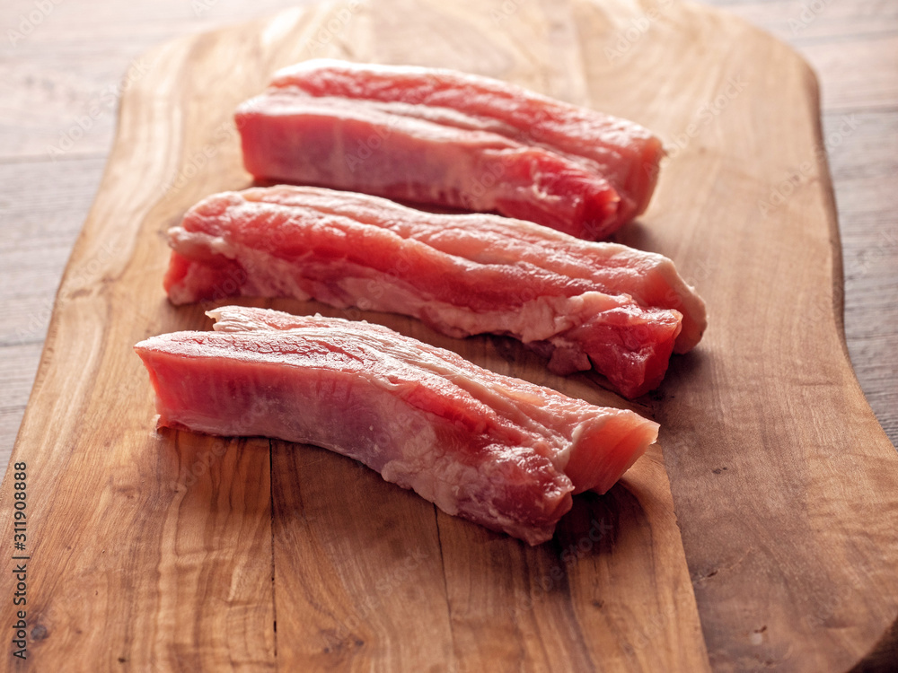 Three raw pork belly slices on a wooden cutting board, Dark wood table surface. Three raw pork belly slices on a wooden cutting board, Dark wood table surface.