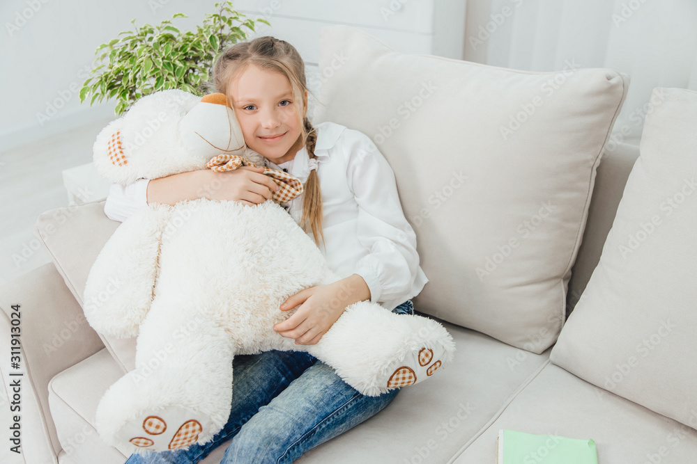 Happy little girl sitting on the sofa, hugging a big teddy-bear ...