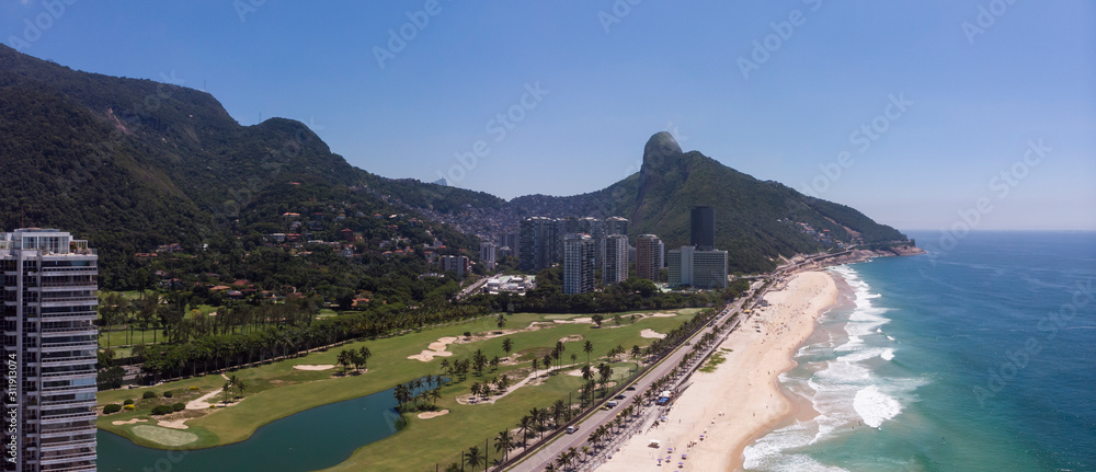 Aerial panorama of coastal São Conrado golf course with high rise building and beach in the foreground and Two Brothers mountain and favela shantytown Rocinha in the background against a blue sky