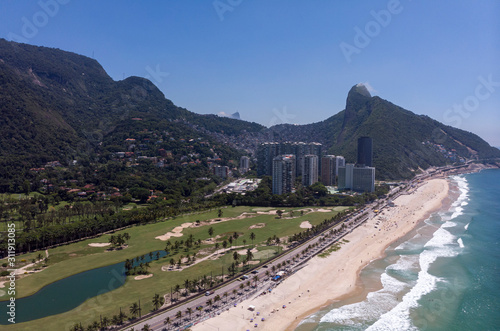 Aerial panorama of coastal São Conrado golf course with beach in the foreground and Two Brothers mountain and Corcovado mountain behind shantytown Rocinha in the background against a blue sky
