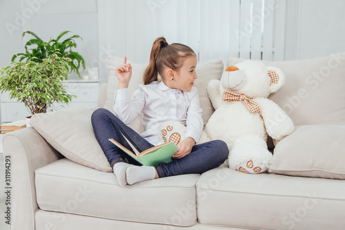 Nice little girl sitting on the sofa with teddy-bear near her, reading fairy-tale, pointing her finger up, saying something important to her soft friend. photo