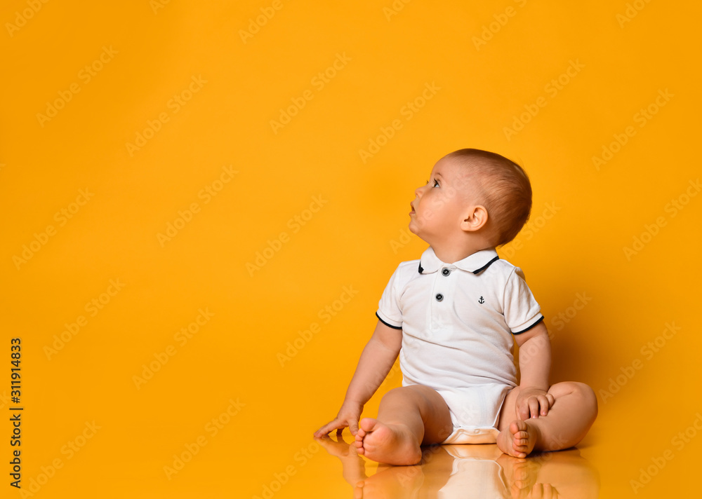 Infant baby boy toddler is sitting on the floor looking up at free copy space in corner yellow background - obrazy, fototapety, plakaty 