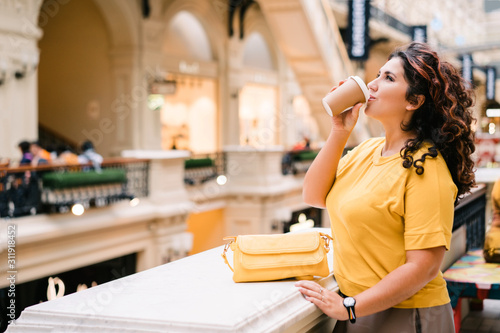 Beautiful cheerful curly red-haired woman in a bright yellow sweater with a handbag with a coffee cup to go in a large shopping center looking at the clock in surprise. Big Sales Shopping