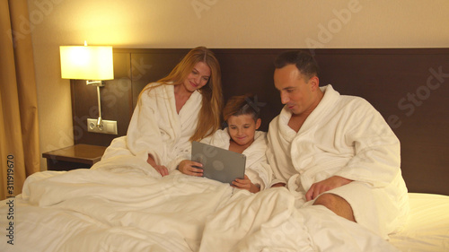 Parents and little son in sit on bed and watch something on tablet in hotel room