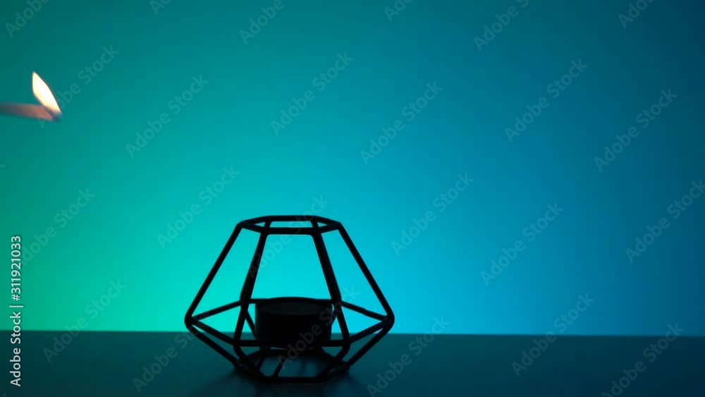 Hand burning candle in black polygonal stand, green and blue background. Relax, spa, Halloween concept. Close-up, copy space Hand burning candle in black polygonal stand, green and blue background. Relax, spa, Halloween concept. Close-up, copy space