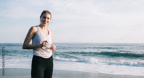 Young smiling sportswoman with water bottle standing on sea beach