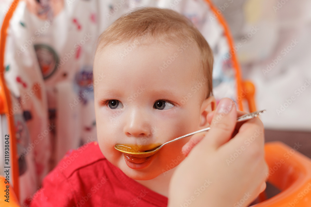 a small child eats with a spoon and sits and holds the edge of the feeding table in the nursery, grimy and stained with food, baby food concept, complementary foods, the first spoon a small child eats with a spoon and sits and holds the edge of the feeding table in the nursery, grimy and stained with food, baby food concept, complementary foods, the first spoon