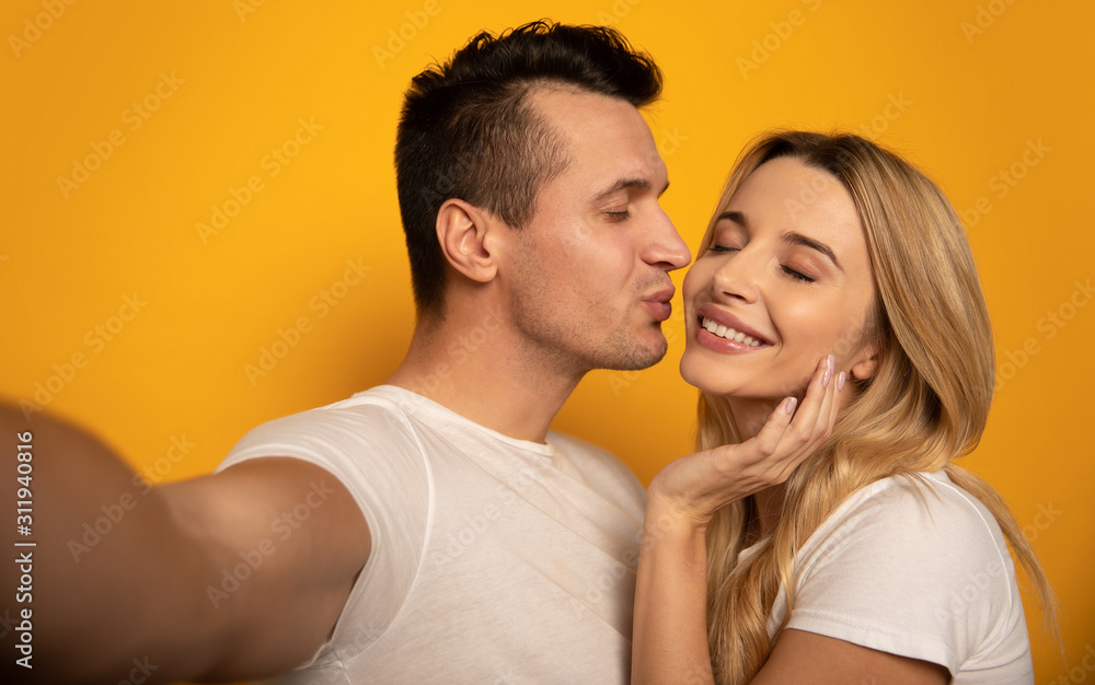 I love you! Close-up photo of an alluring girl with fair hair and a handsome man, who is kissing his girlfriend in a cheek while taking a selfie with her.