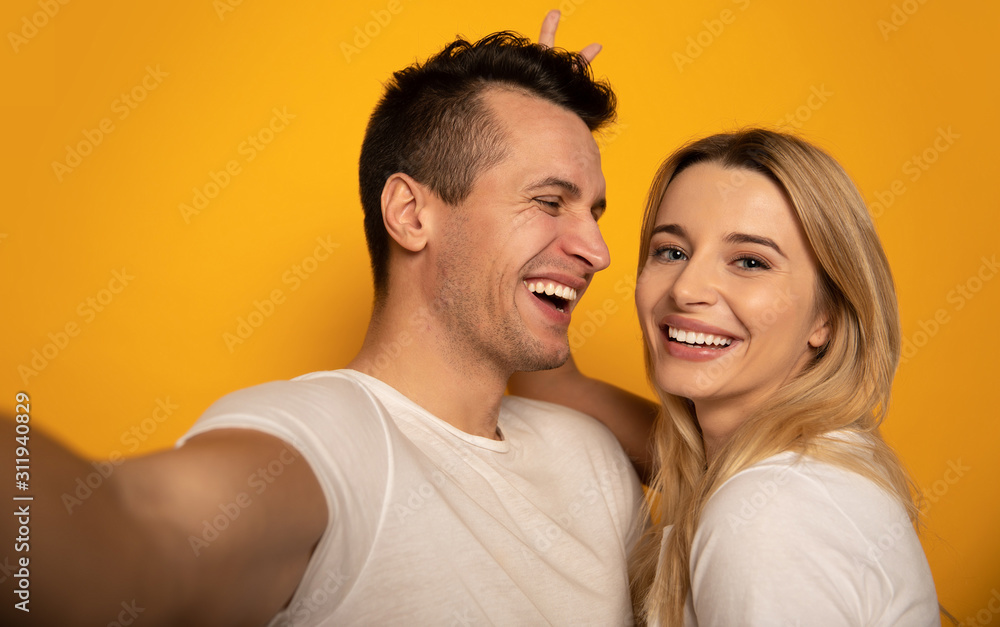 Crazy lovely couple. Close-up photo of a cute girl and a cheerful man, who are laughing while making a joint selfie, and a girl is showing a v-sign behind his head.