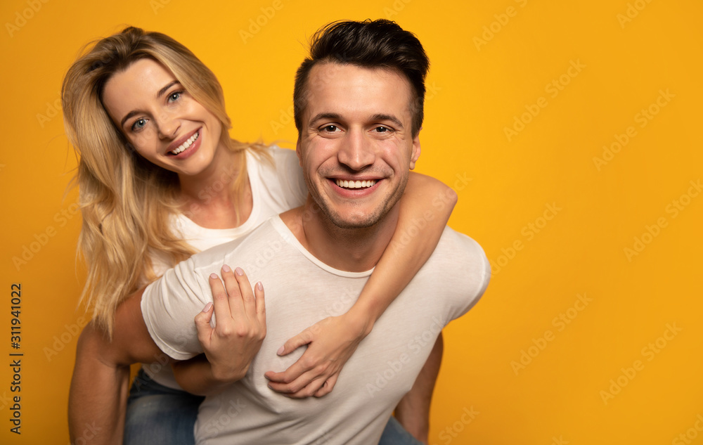 A good company. Close-up photo of an attractive man with raven black hair, who is holding his beautiful girlfriend on his back and looking in the camera with a broad smile. A good company. Close-up photo of an attractive man with raven black hair, who is holding his beautiful girlfriend on his back and looking in the camera with a broad smile.