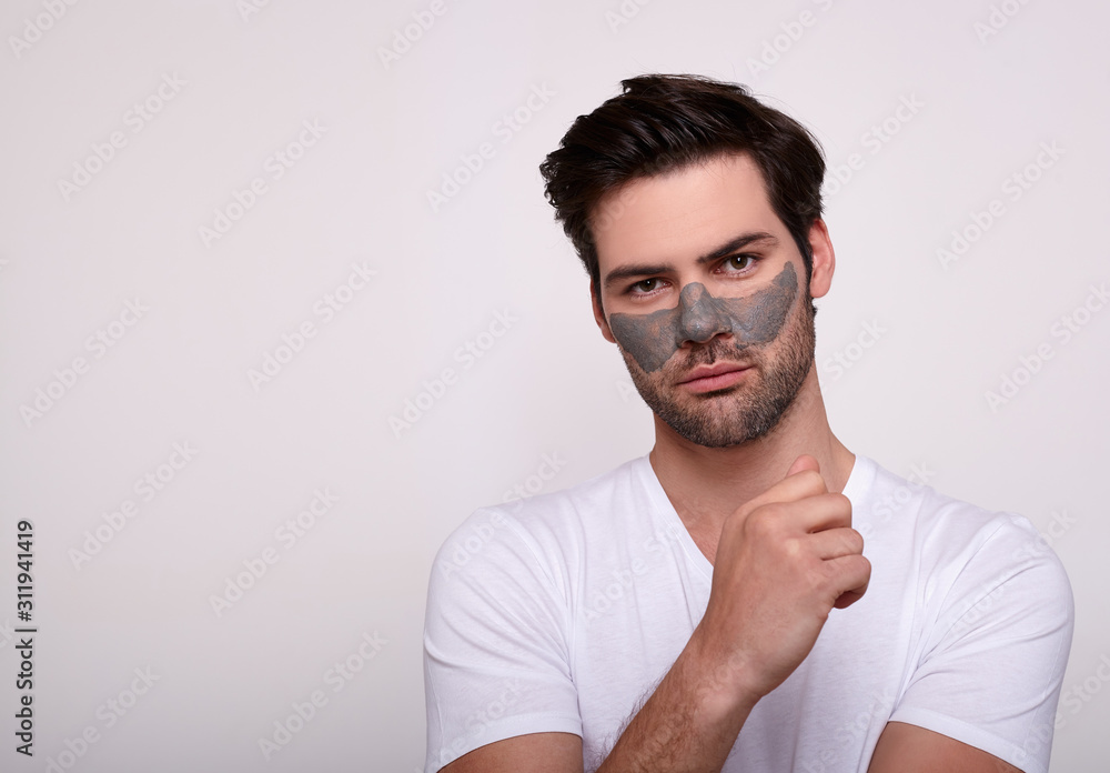 A calm young handsome man in a dark clay face mask on a white background. A calm young handsome man in a dark clay face mask on a white background.