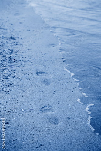 Closeup of feet traces footprints on sand beach by sea ocean water. Toned with classic blue 2020 colour. Natural eco environmental background texture.