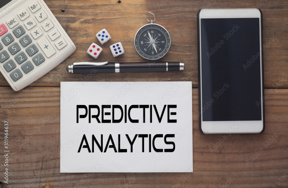 Predictive Analytics written on paper,Wooden background desk with calculator,dice,compass,smart phone and pen.Top view conceptual.