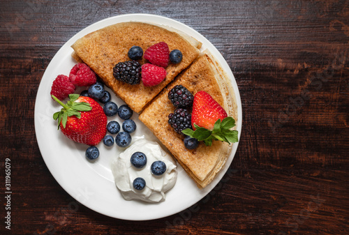 Pancakes with berries and sour cream on a dark background. Food background. Healthy and delicious breakfast. Sweet pancakes in a white plate on a wooden table. Top view