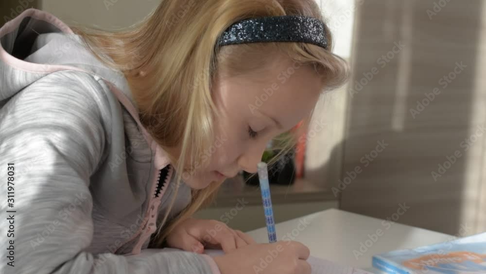 Blonde girl of 8 year old learning homework, girl doing her lessons, schoolgirl studying at home on white table. Concept back to school. Side view, close up. Blonde girl of 8 year old learning homework, girl doing her lessons, schoolgirl studying at home on white table. Concept back to school. Side view, close up.