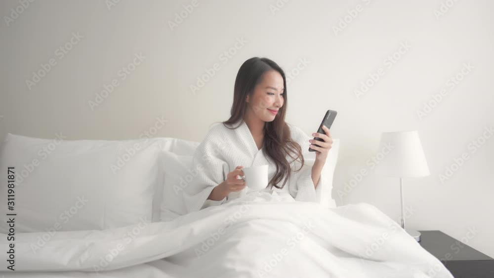 Young woman wearing wearing a dressing gown, holding a cup of coffee and using her mobile phone while lying in her bed in the morning. Young woman wearing wearing a dressing gown, holding a cup of coffee and using her mobile phone while lying in her bed in the morning.