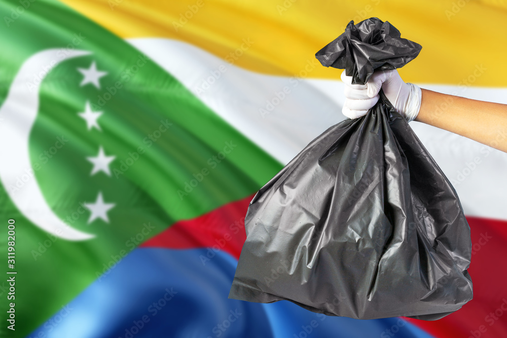 Comoros environmental protection concept. The male hand holding a garbage bag on national flag background. Ecological and recycling theme with copy space.