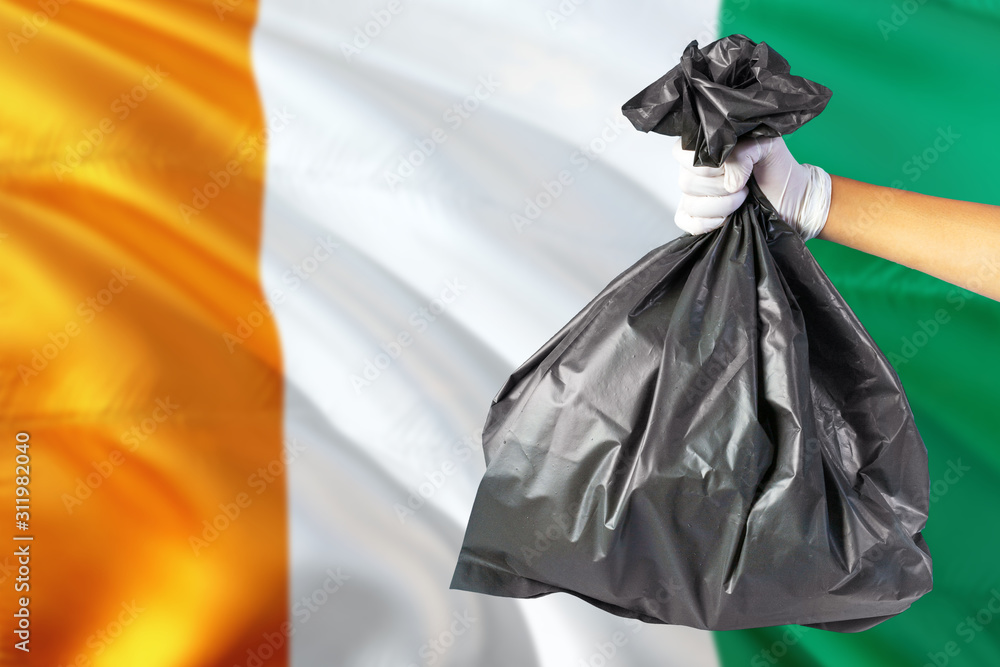 Cote D'Ivoire environmental protection concept. The male hand holding a garbage bag on national flag background. Ecological and recycling theme with copy space.