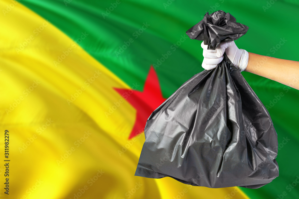 French Guiana environmental protection concept. The male hand holding a garbage bag on national flag background. Ecological and recycling theme with copy space.