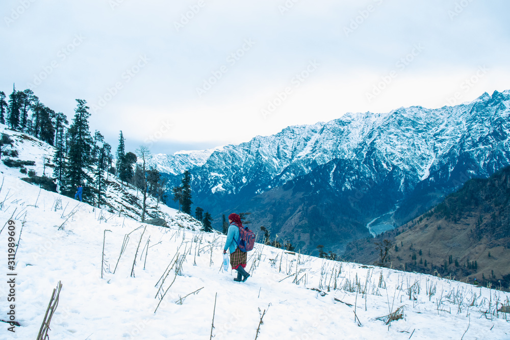 Woman walking on the snow carrying bag and tea kettel to serve tourist hot tea in winter tourist season with beautiful snow mountain landscape in the background, holiday destination, kashmir, India.