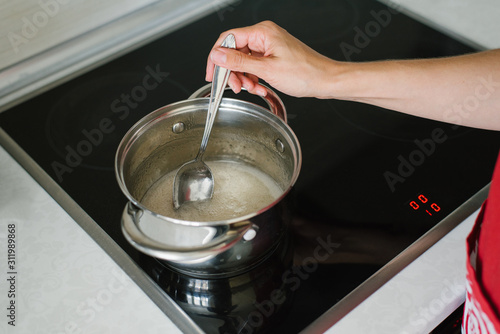 A woman's hand stirs the sugar syrup liquid in a saucepan on an electric stove with a tablespoon