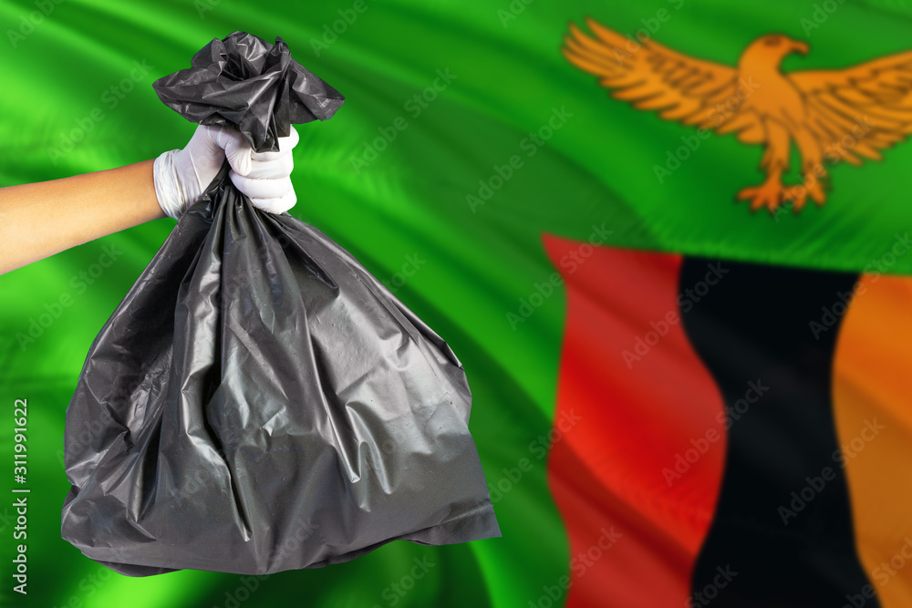 Zambia environmental protection concept. The male hand holding a garbage bag on national flag background. Ecological and recycling theme with copy space.