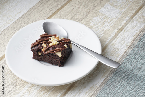 A moist dark chocolate brownie with mix nut on top served on white round plate with silver spoon.