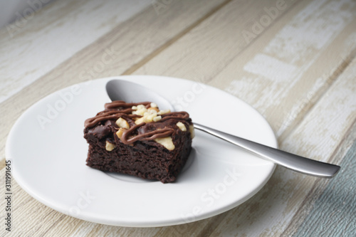 A moist dark chocolate brownie with mix nut on top served on white round plate with silver spoon.