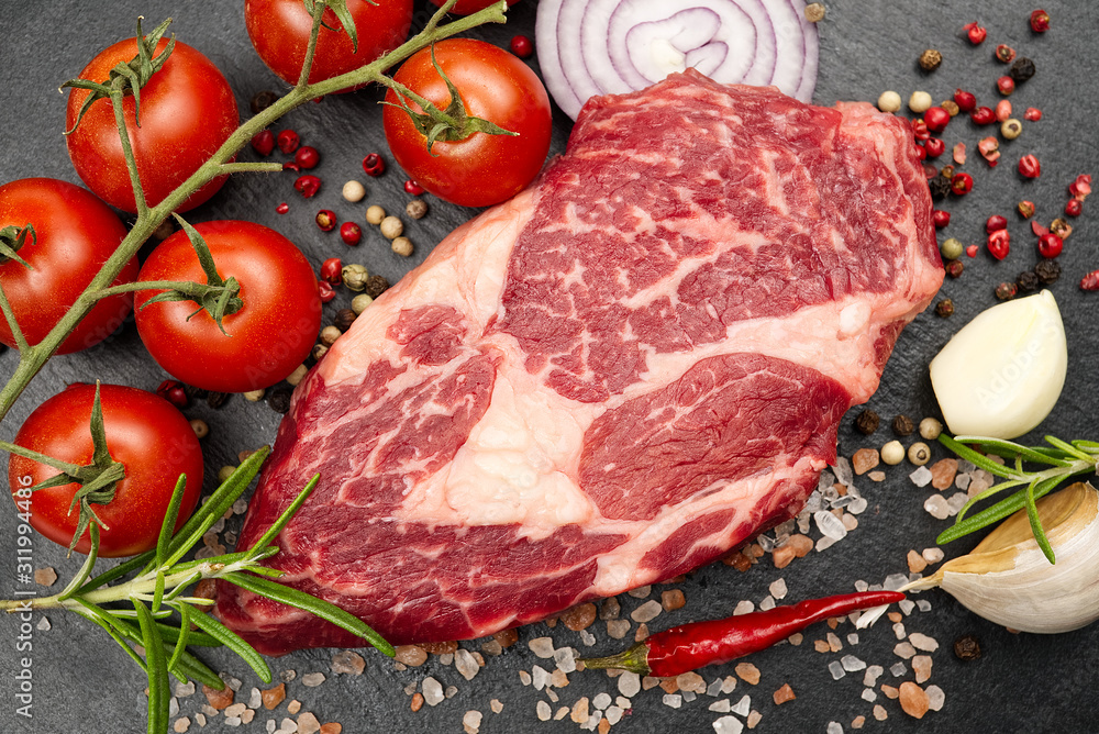 Raw rib eye steak with spices and vegetables. Ingredients for restaurant meal. Fresh meat, salt, rosemary, thyme, chilli, cherry tomatoes, garlic on black stone. Food background. Raw rib eye steak with spices and vegetables. Ingredients for restaurant meal. Fresh meat, salt, rosemary, thyme, chilli, cherry tomatoes, garlic on black stone. Food background.