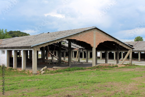 Densely built wide large hangar buildings with missing destroyed red brick support walls and unusual bent roofs surrounded with paved area mixed with grass and trees at abandoned military complex © hecos