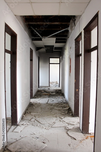 Fototapeta Naklejka Na Ścianę i Meble -  Narrow corridor with broken ceiling tiles and missing doors surrounded with cracked walls and destroyed floor tiles at abandoned building on warm sunny summer day