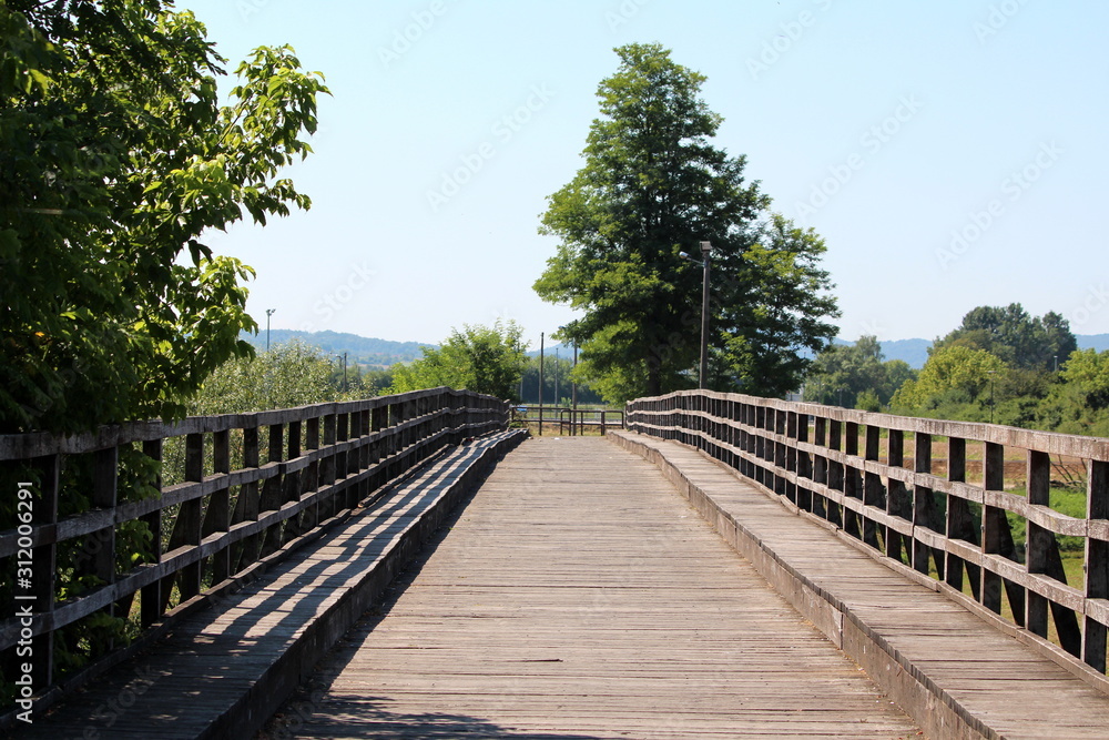 Old massive dilapidated pedestrian wooden bridge with wooden handrails surrounded with tall dense trees and clear blue sky in background