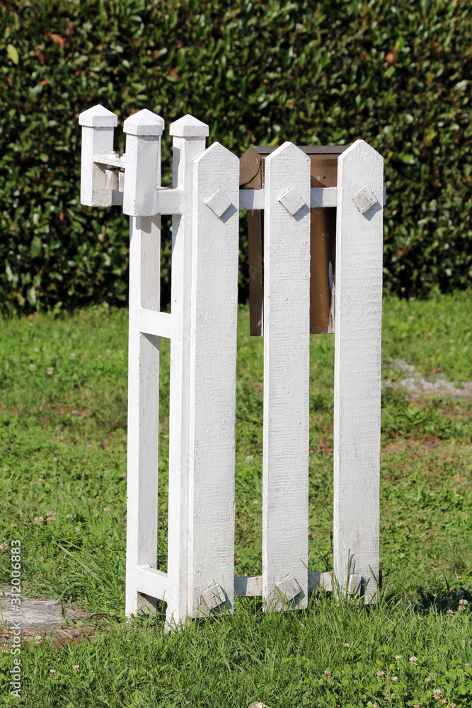 Part of newly made white wooden picket fence with mounted brown metal mailbox surrounded with uncut grass and dark green hedge in background