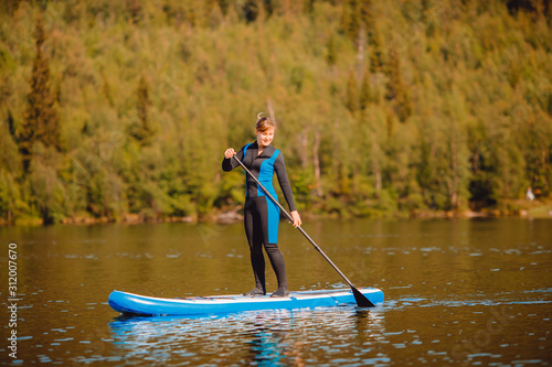 Girl in thermo clothing rowing oar on sup board blue lake water paddleboard background of forest © Parilov