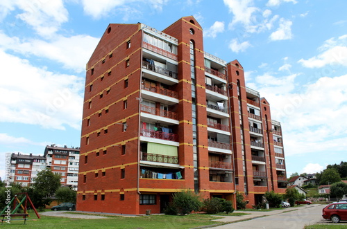 Vintage large red brick apartment building with long balconies with red metal fences surrounded with grass and paved parking lot in front on cloudy blue sky background
