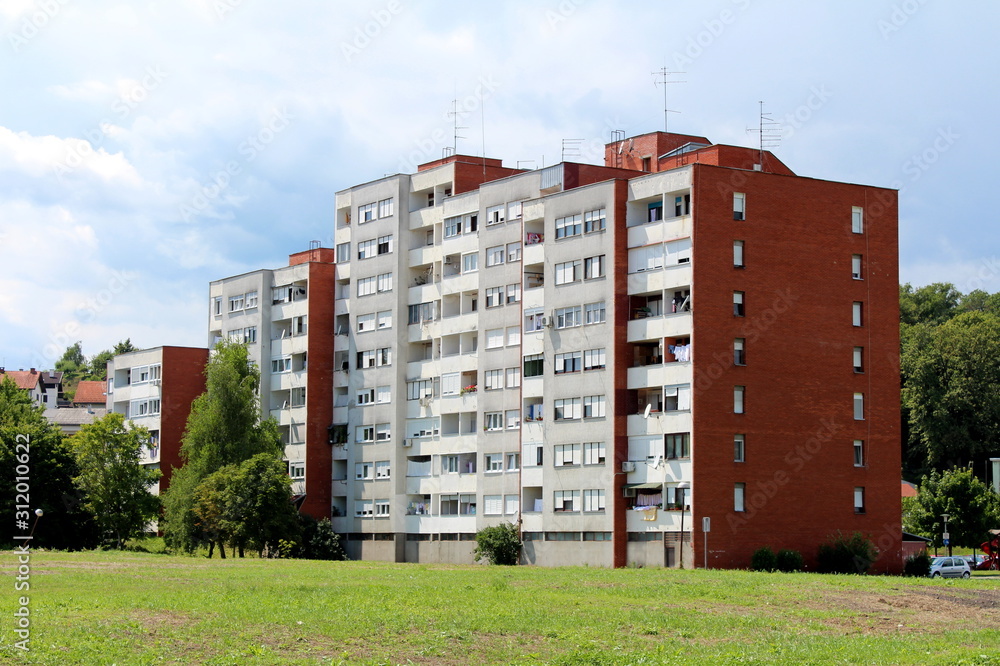 White apartment building with red building block sidewalls and multiple balconies surrounded with grass and tall trees on cloudy blue sky background