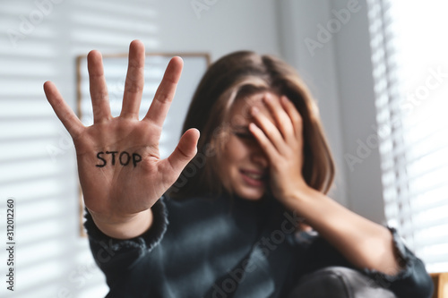 Crying young woman showing palm with word STOP indoors, focus on hand. Domestic violence concept