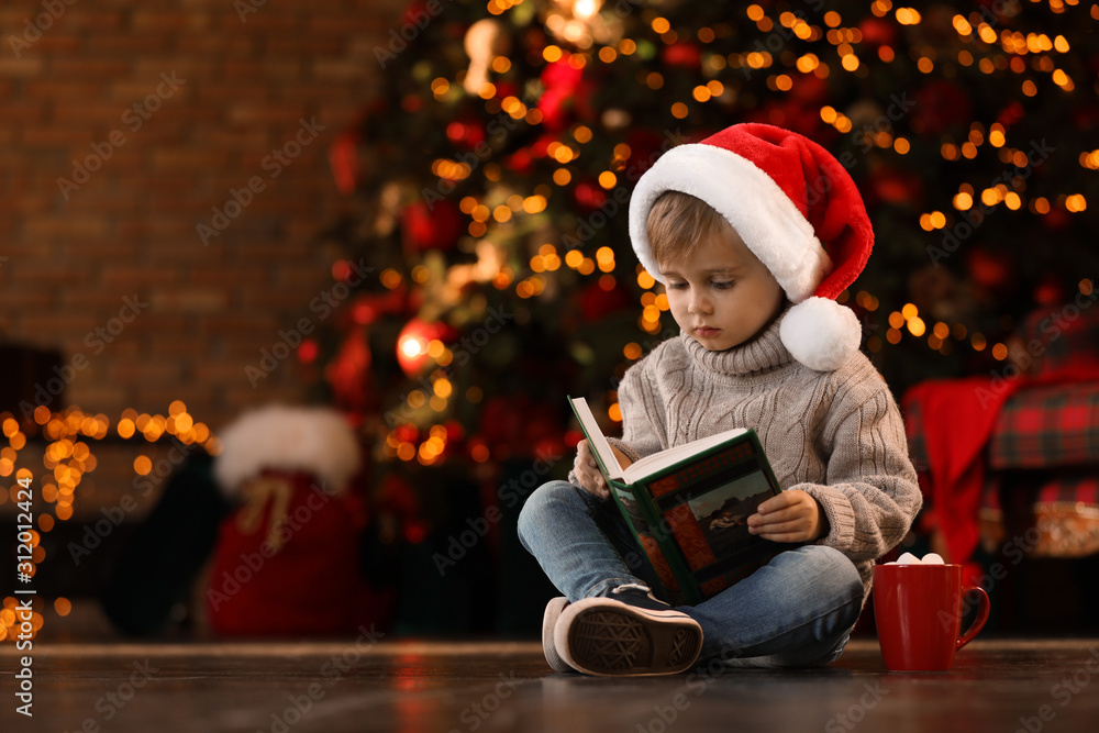 Little boy in Santa Claus cap reading book near Christmas tree at home Little boy in Santa Claus cap reading book near Christmas tree at home