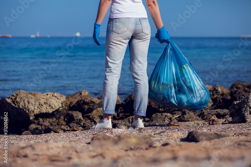 Woman volunteer with big blue bag collecting garbage on beach. Environmental pollution concept