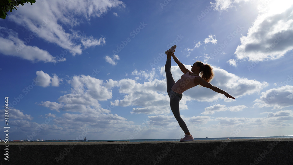 Flexible athletic girl performs dance pose elements on a background of bright blue sky with white clouds. Curly-haired gymnast does the splits. Dancer, ballerina, athlete