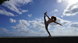 Flexible athletic girl performs dance pose elements on a background of bright blue sky with white clouds. Curly-haired gymnast does the splits. Dancer, ballerina, athlete